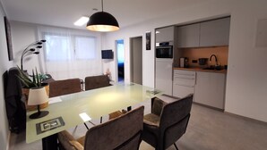 Une cuisine moderne dotée d’un four et d’un micro-ondes encastrés, un coin repas avec une table et des chaises en verre, et un téléviseur fixé au mur.