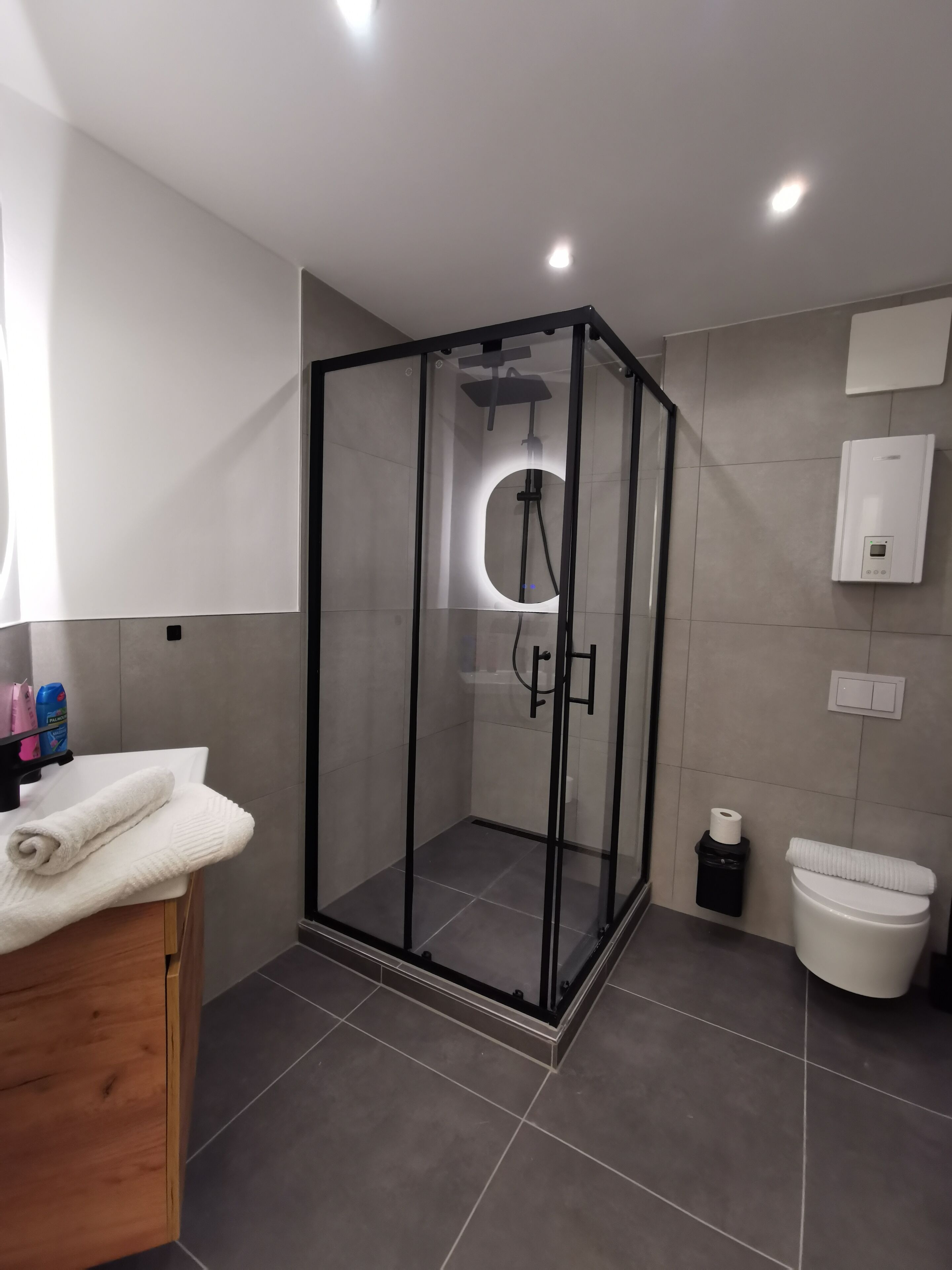 A modern bathroom with a glass-enclosed shower, a wooden vanity, a toilet, and a wall-mounted heater.