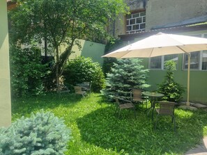 Een terras met een tafel en stoelen onder een grote parasol, omgeven door weelderig groen en bomen.