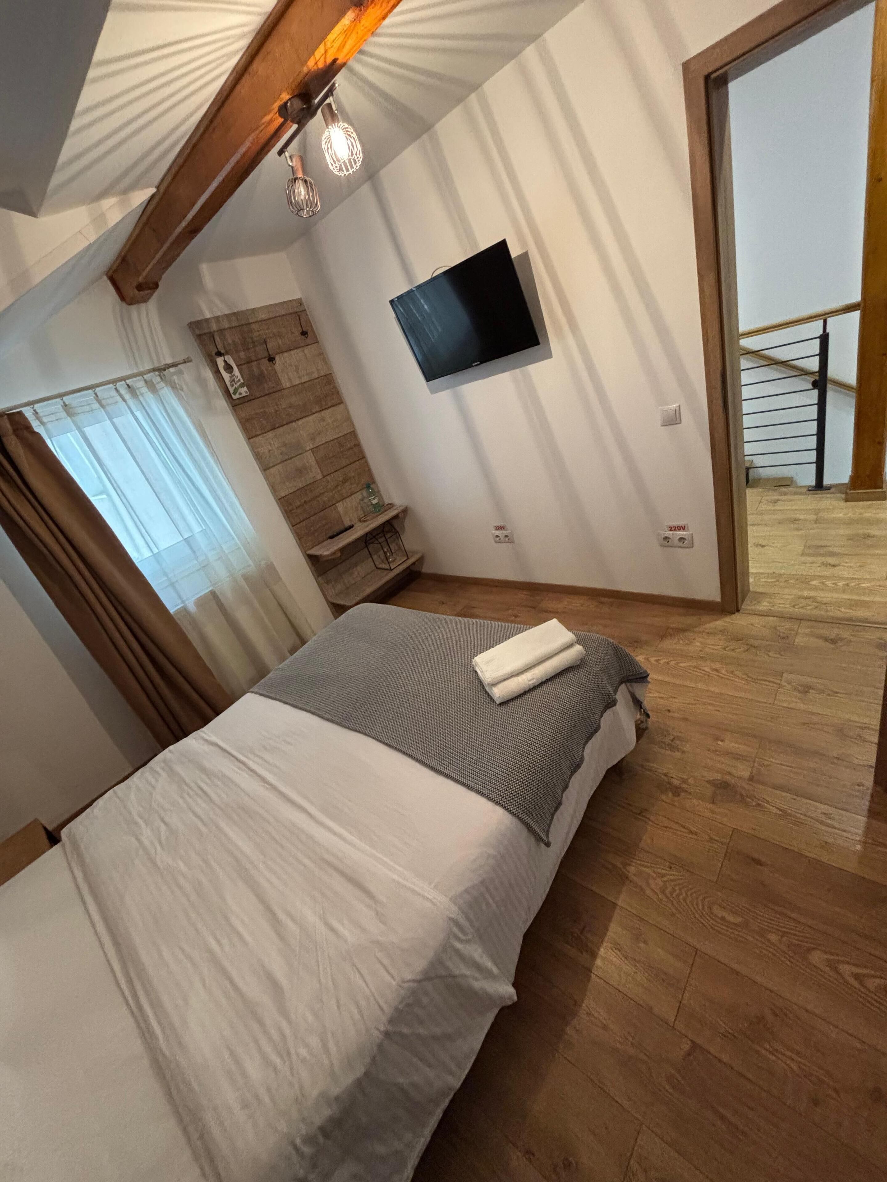 A bedroom with a bed, a TV mounted on the wall, a wooden staircase, and a view of a balcony.
