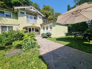 Un edificio de dos pisos con un balcón, un sendero de piedra que lleva a la entrada y mobiliario exterior.
