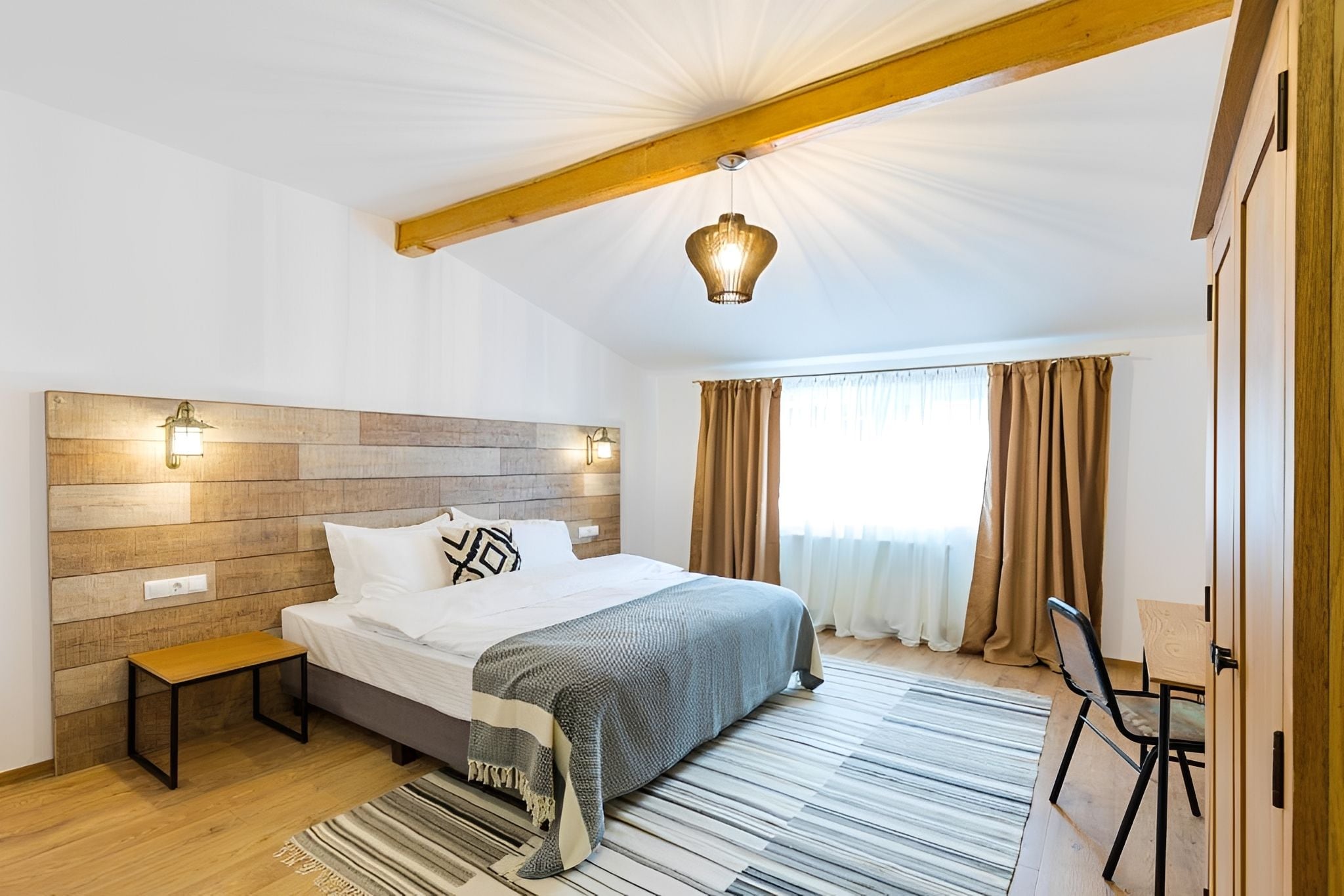 Un dormitorio moderno con cabecera de madera, cama con sábanas blancas, un banco, un escritorio y un amplio ventanal con cortinas.