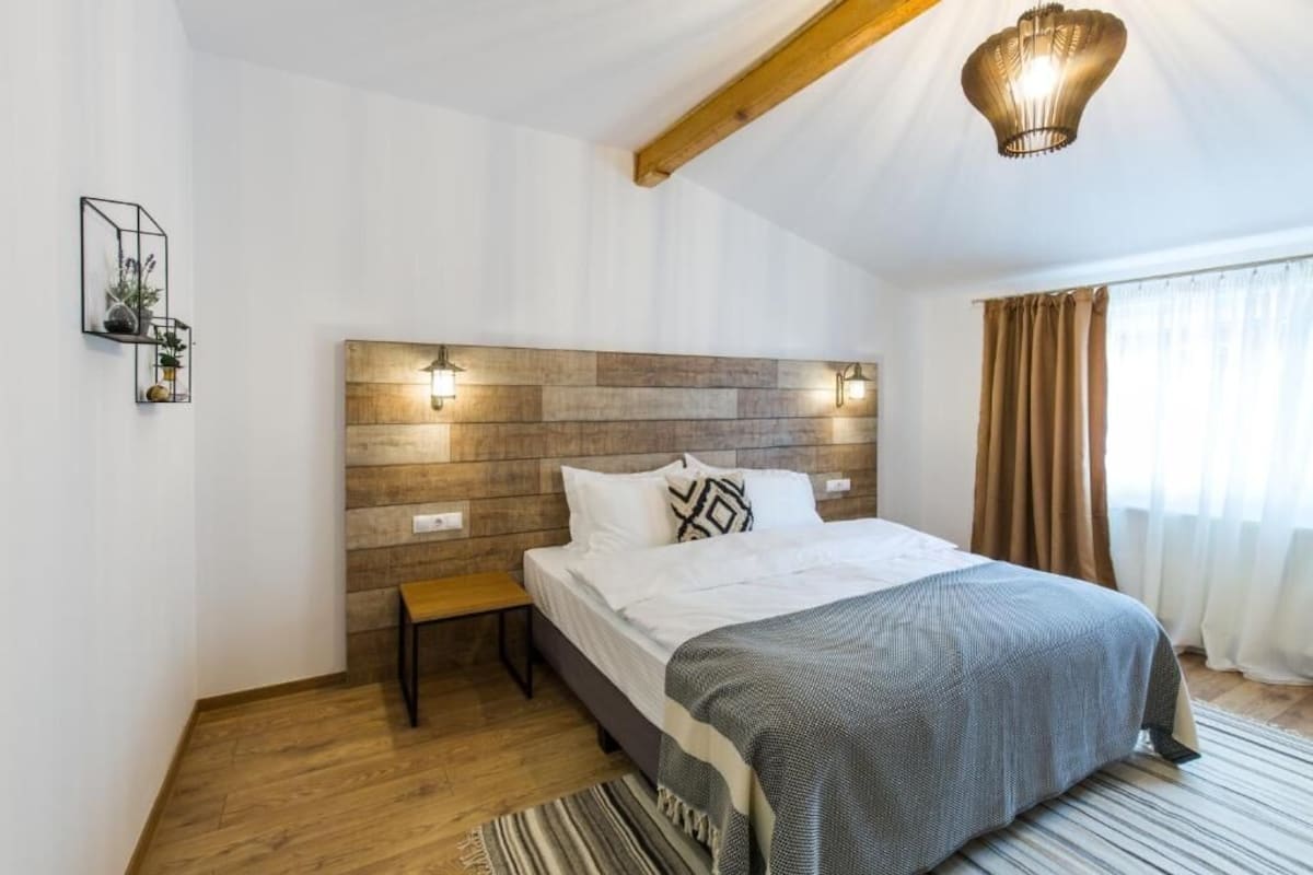 Ein Schlafzimmer mit einem hölzernen Kopfteil, einem Bett mit grauer Decke, einem Nachttisch und einer Pendelleuchte.