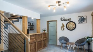 Un intérieur moderne avec un escalier en bois, une pancarte « Bienvenue », une horloge murale et deux chaises grises.
