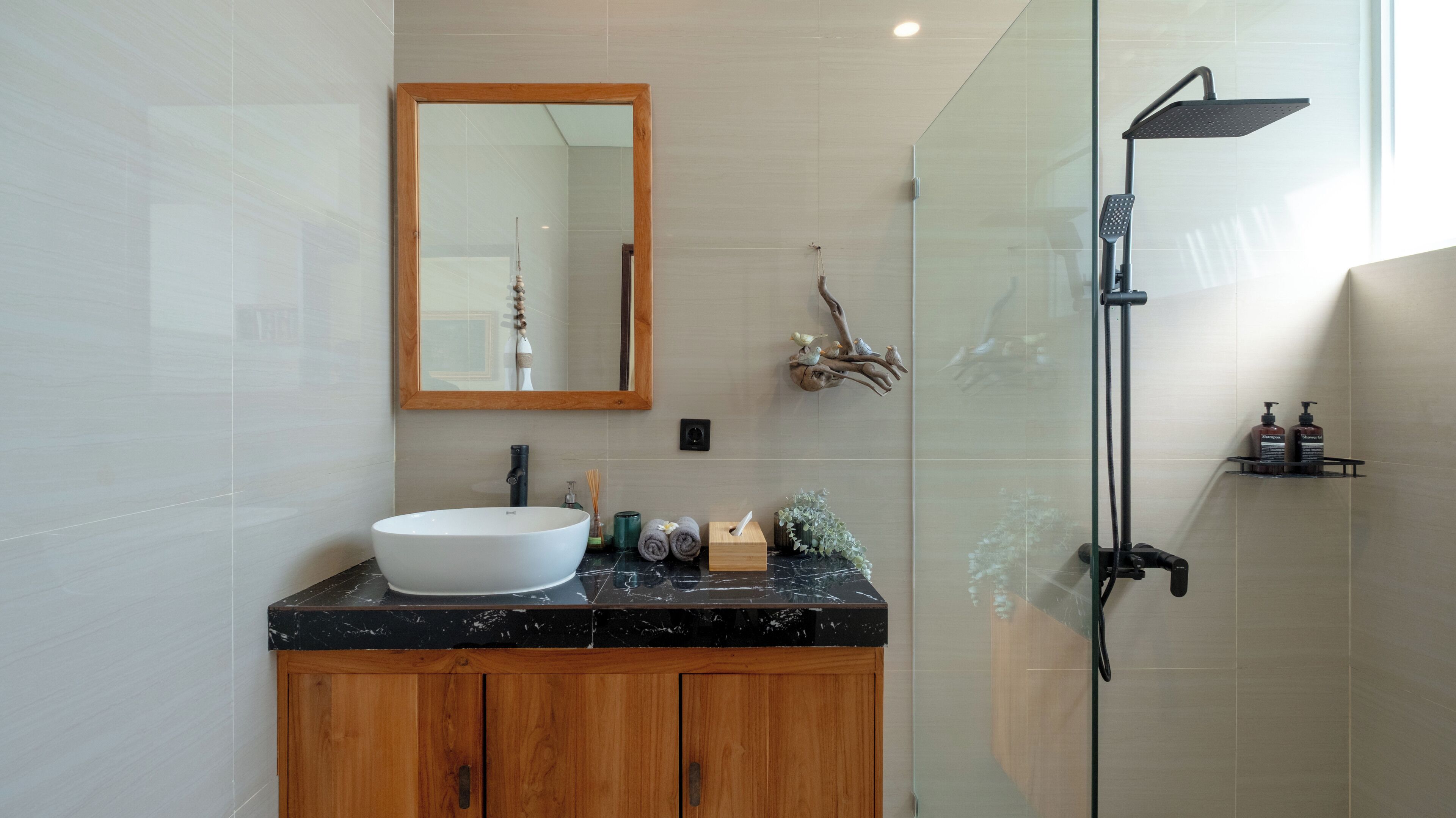 A modern bathroom with a large white sink, wooden cabinets, a mirror, a glass shower enclosure, and a small shelf with toiletries.