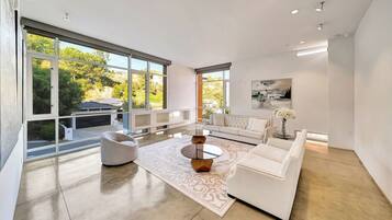 A modern living room with a white sofa, a glass coffee table, and a large window offering a view of greenery and a building.