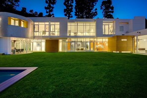 A modern two-story house with a large front yard, a swimming pool, and a well-lit interior.