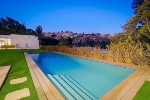A swimming pool with a wooden deck and artificial grass on one side, surrounded by dry vegetation and hills in the background.