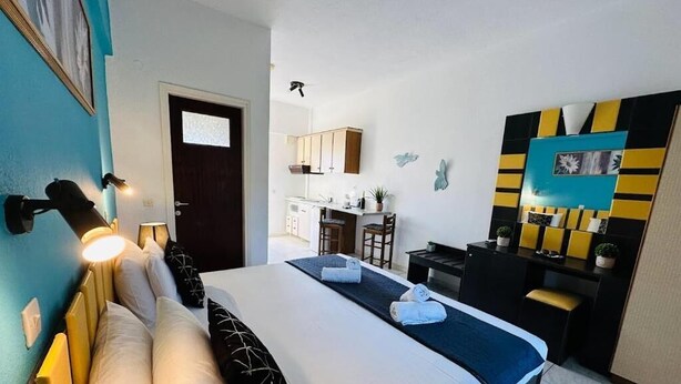 A hotel room with a bed, bedside tables, a desk, and a chair. There is a door leading to a kitchenette and a bathroom. - Marina studios (Corfu)