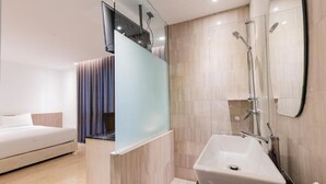 Habitación de hotel moderna con una cama grande, un separador de vidrio, un baño con lavamanos y ducha, y un espejo amplio.
