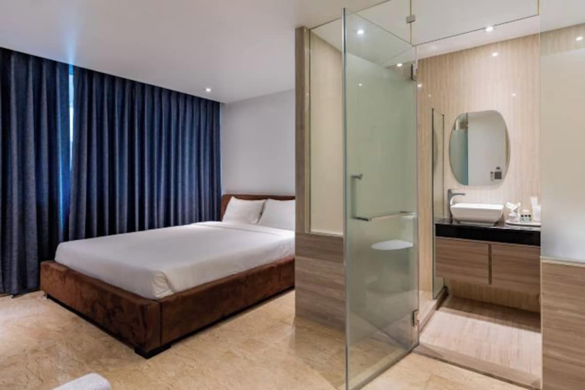 A modern hotel room with a large bed, a glass-enclosed shower, and a wooden vanity with a mirror.