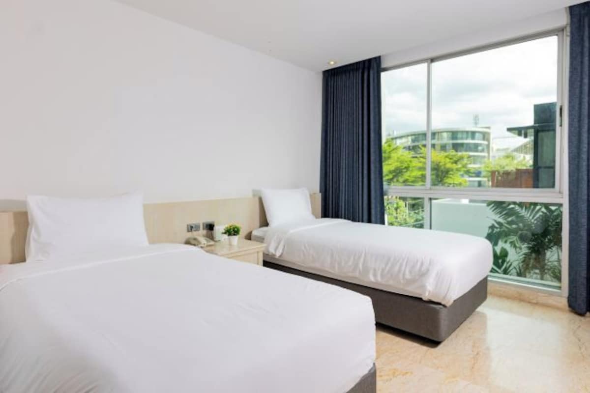 A hotel room with two beds, a large window with curtains, and a view of buildings and greenery outside.