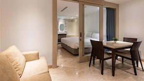 Una habitación de hotel moderna con una cama, una mesa de comedor con sillas, un sofá y un baño visible a través de una puerta de vidrio.