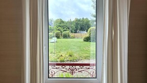 Une vue à travers une fenêtre ornée de rideaux blancs, dévoilant un jardin bien entretenu avec une pelouse, des arbustes et un chemin.