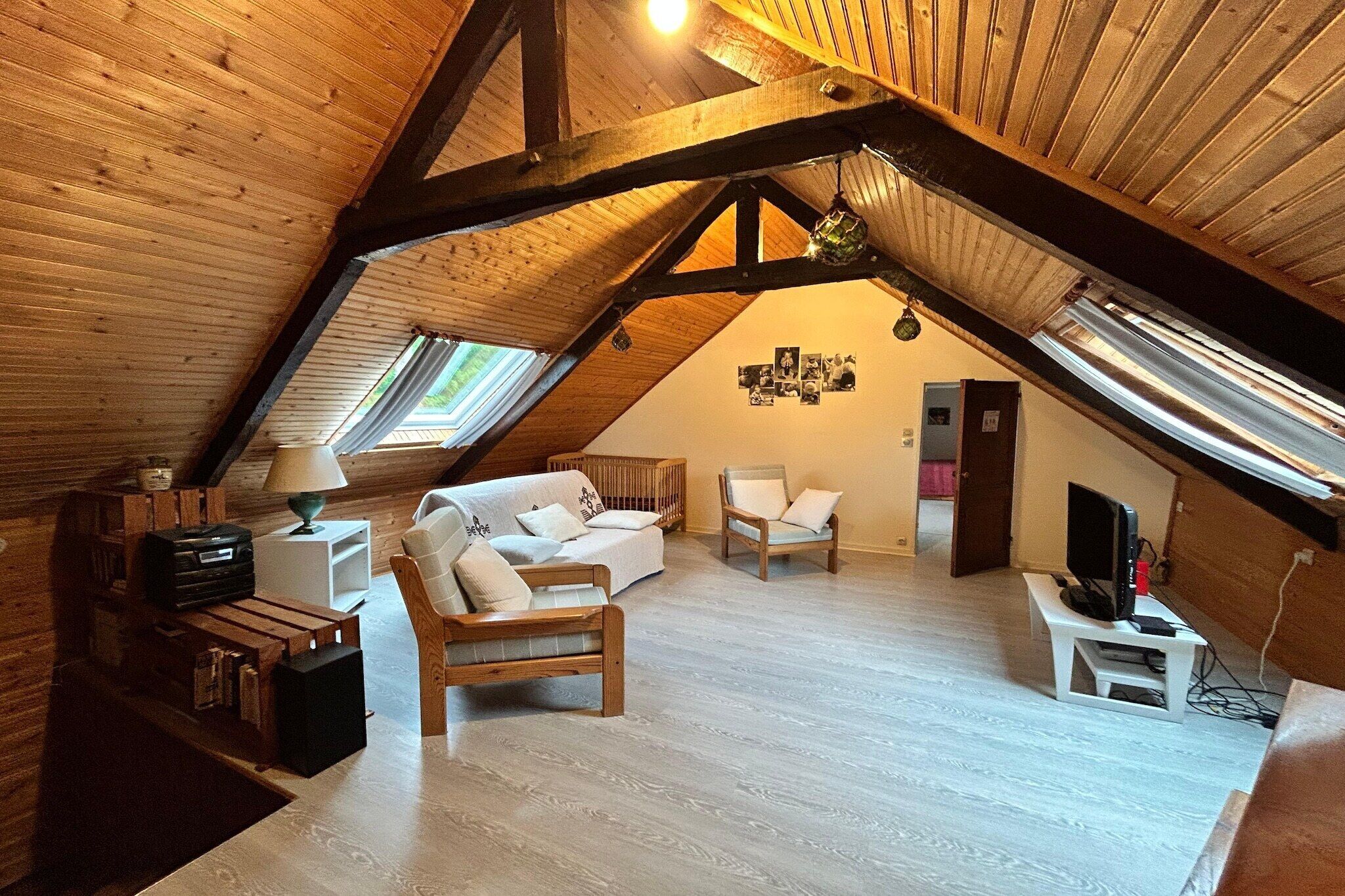 Quarto de sótão aconchegante, com vigas de madeira, um sofá branco, uma mesa pequena com abajur e uma televisão em um suporte.