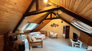 Une pièce mansardée chaleureuse avec des poutres apparentes, un canapé blanc, une petite table avec une lampe et un téléviseur posé sur un meuble.