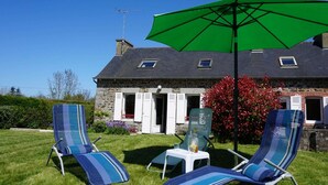 Une maison en pierre avec un toit en pente, un parasol vert et deux fauteuils de jardin à rayures.