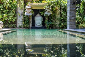 Ein friedlicher Poolbereich mit einer Buddha-Statue, umgeben von üppiger Vegetation und steinernen Säulen.