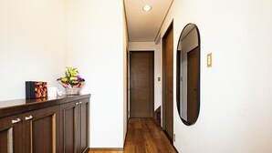 A hallway with wooden flooring, a tiled area at the end, a dark wooden cabinet, a vase with flowers, and a mirror on the wall. - HITO (Fukuoka)