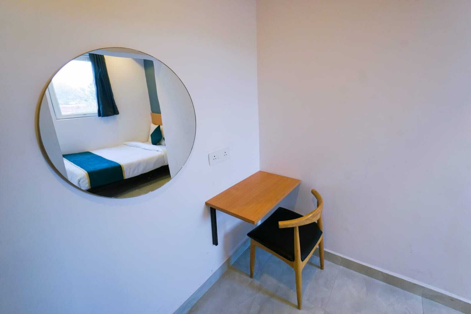 A small room with a round mirror, a wooden table, a black chair, and a bed with blue and white bedding.