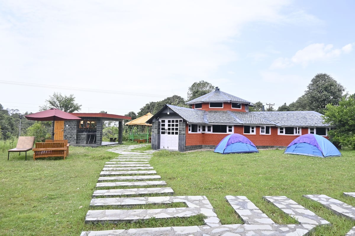 A rustic lodge with a stone pathway leading to a wooden bench and a small shelter. Two tents are set up on the lawn.