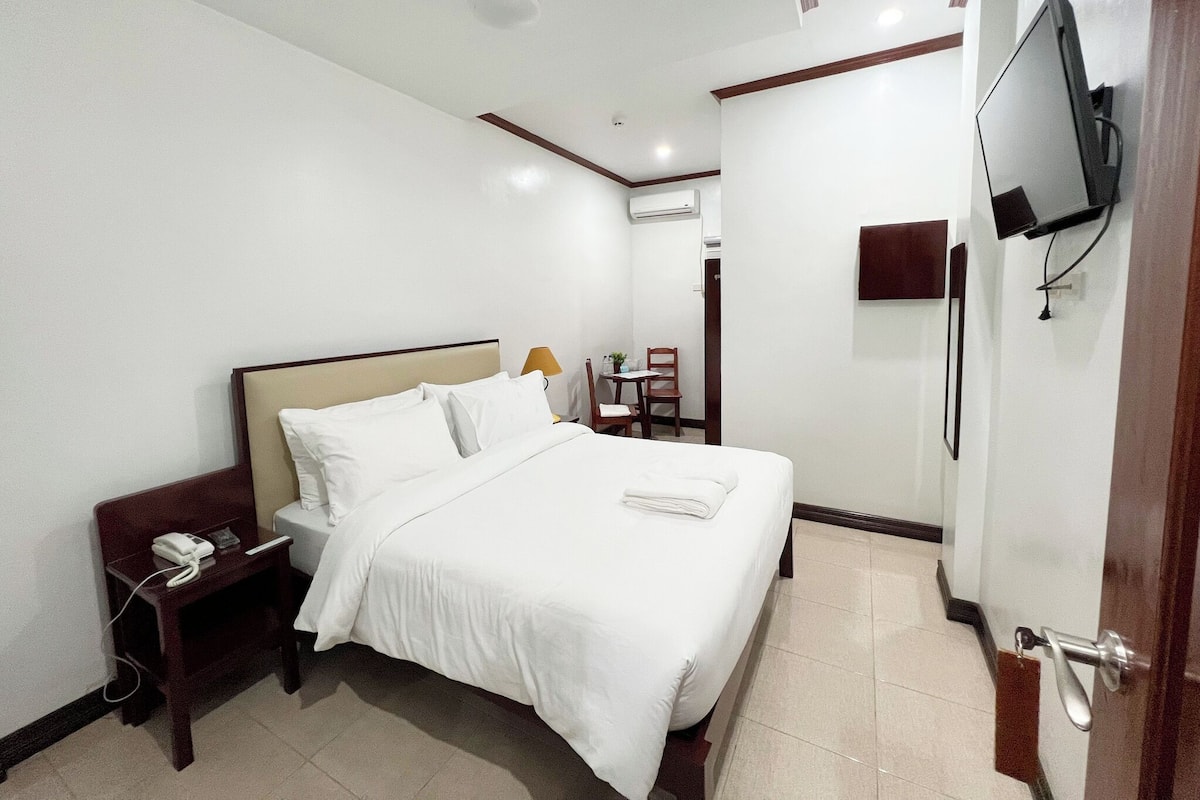 Habitación de hotel con cama, mesita de noche, lámpara, televisor y una pequeña mesa con una silla.