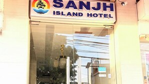 La entrada del Hotel Sanjh Island, con un frente de vidrio transparente, un toldo rojo y un letrero que indica que el hotel está abierto.