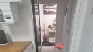 Ein schmaler Flur mit einem schwarzen, flauschigen Teppich am Ende. Links ist eine kleine Küchenzeile mit Mikrowelle und Herd zu sehen. Im Hintergrund befinden sich eine Topfpflanze und ein rotes Objekt.