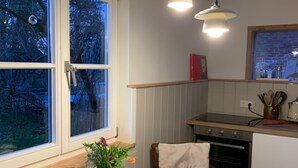 A kitchen with a white table, two pendant lights, a window with a view of trees, and a vase with flowers on the table. - Holiday Home 'Sommerhaus' with Private Terrace, Private Garden and Wi-Fi (Zülow)