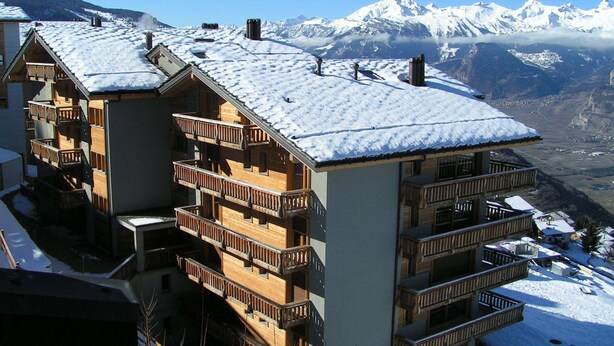 A multi-story building with wooden balconies and a snow-covered roof, set against a mountainous backdrop. - Charming apartment with ski storage (Veysonnaz)