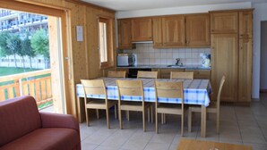 A kitchen with wooden cabinets, a tiled backsplash, a dining table with chairs, and a view of a balcony. - Charming apartment with ski storage (Veysonnaz)