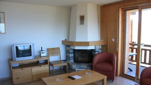 A living room with a stone fireplace, wooden furniture, a television, and a sliding glass door leading outside. - Charming apartment with ski storage (Veysonnaz)