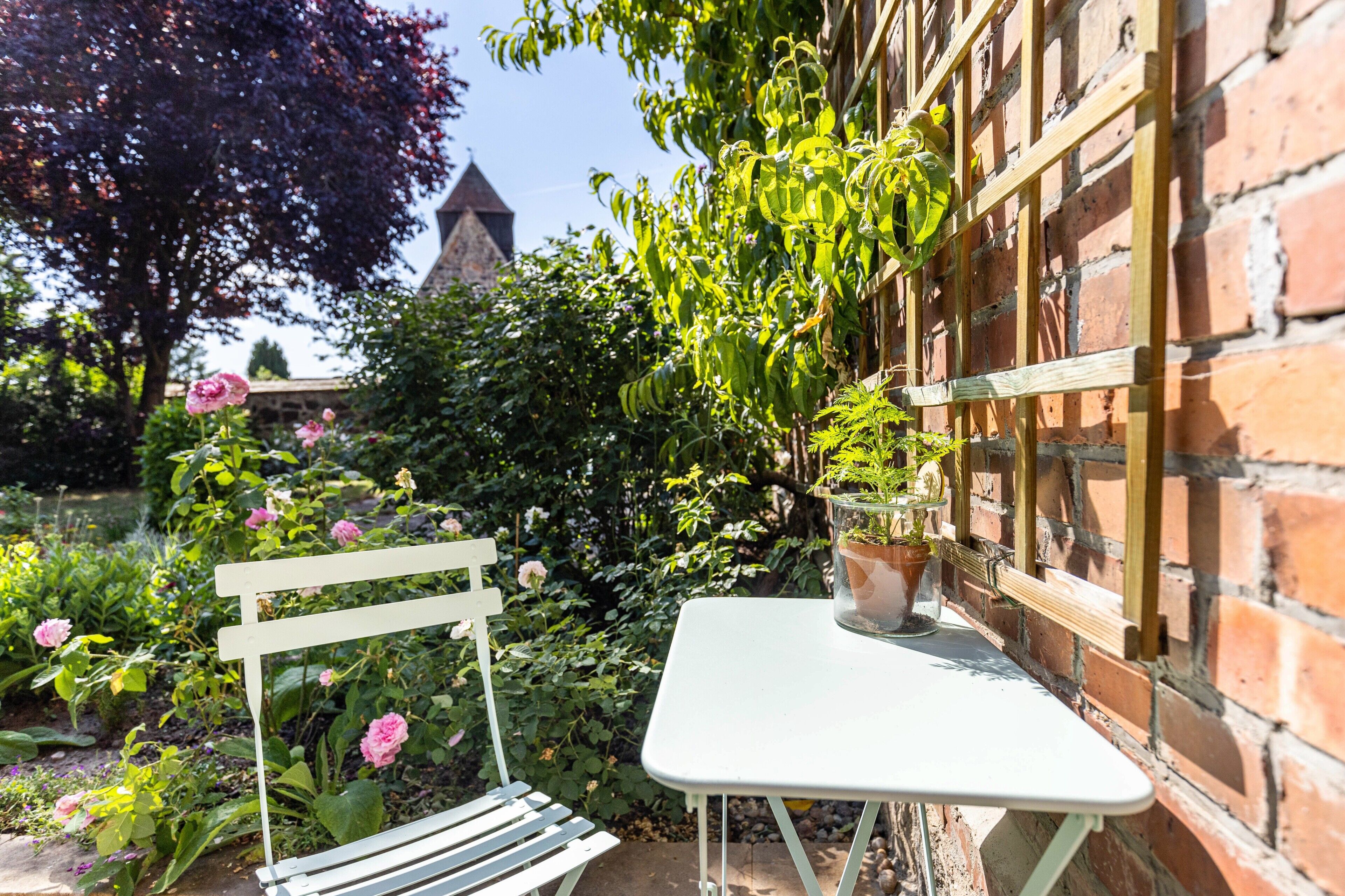 A garden with a white bench and table, a brick wall, and a building with a pointed roof in the background.