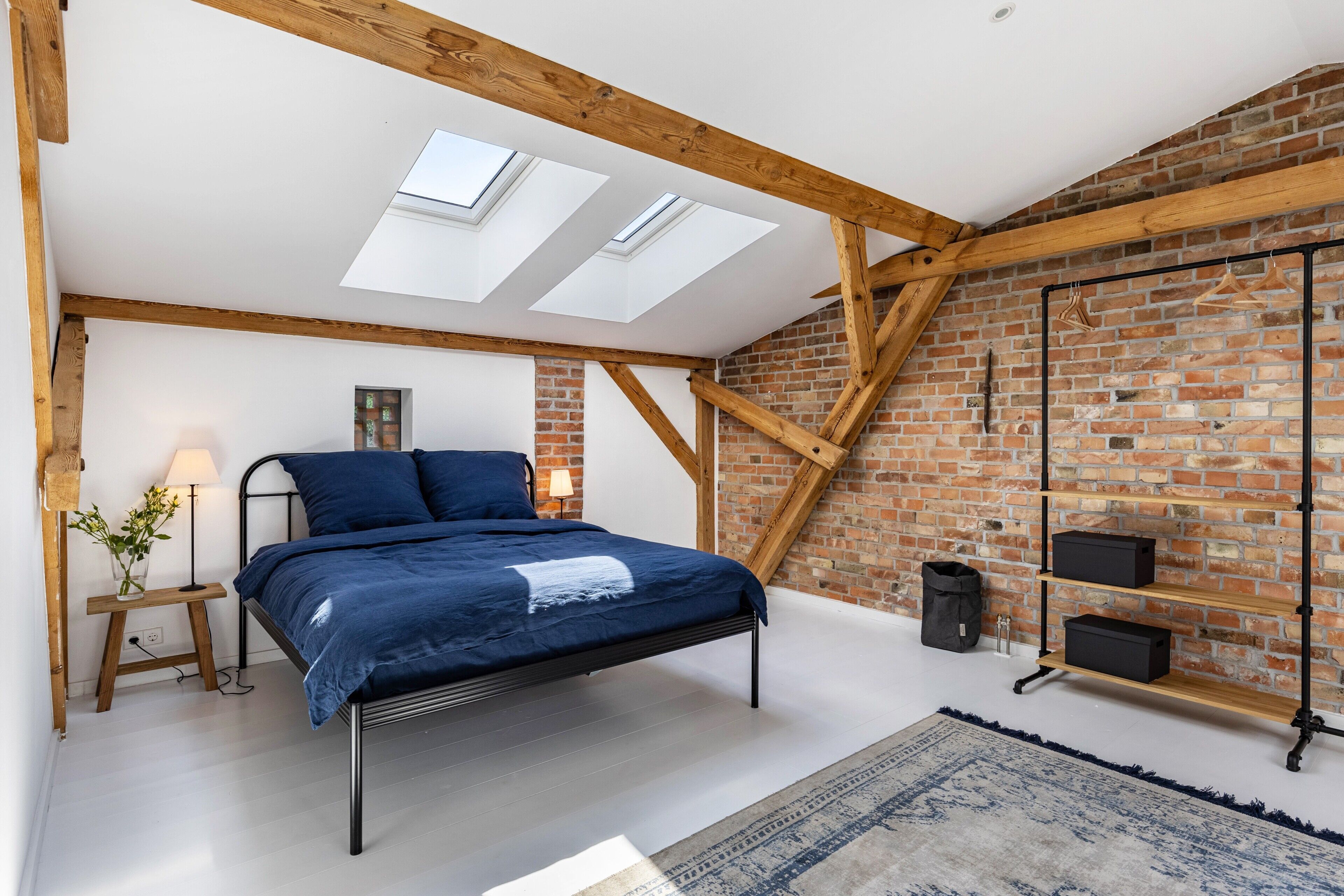 A bedroom with a bed, skylights, a brick wall, a wooden beam ceiling, a bedside table, a lamp, a rug, and a black storage unit.