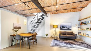 A spacious loft with a high ceiling, exposed brick, a dining area, a sofa, a bookshelf, and a staircase. - Apartment in Former Horse Stable (Angermünde)