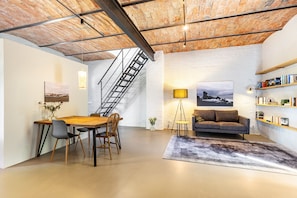 Un loft spacieux avec de hauts plafonds, des murs en briques apparentes, un coin repas, un canapé, une bibliothèque et un escalier.