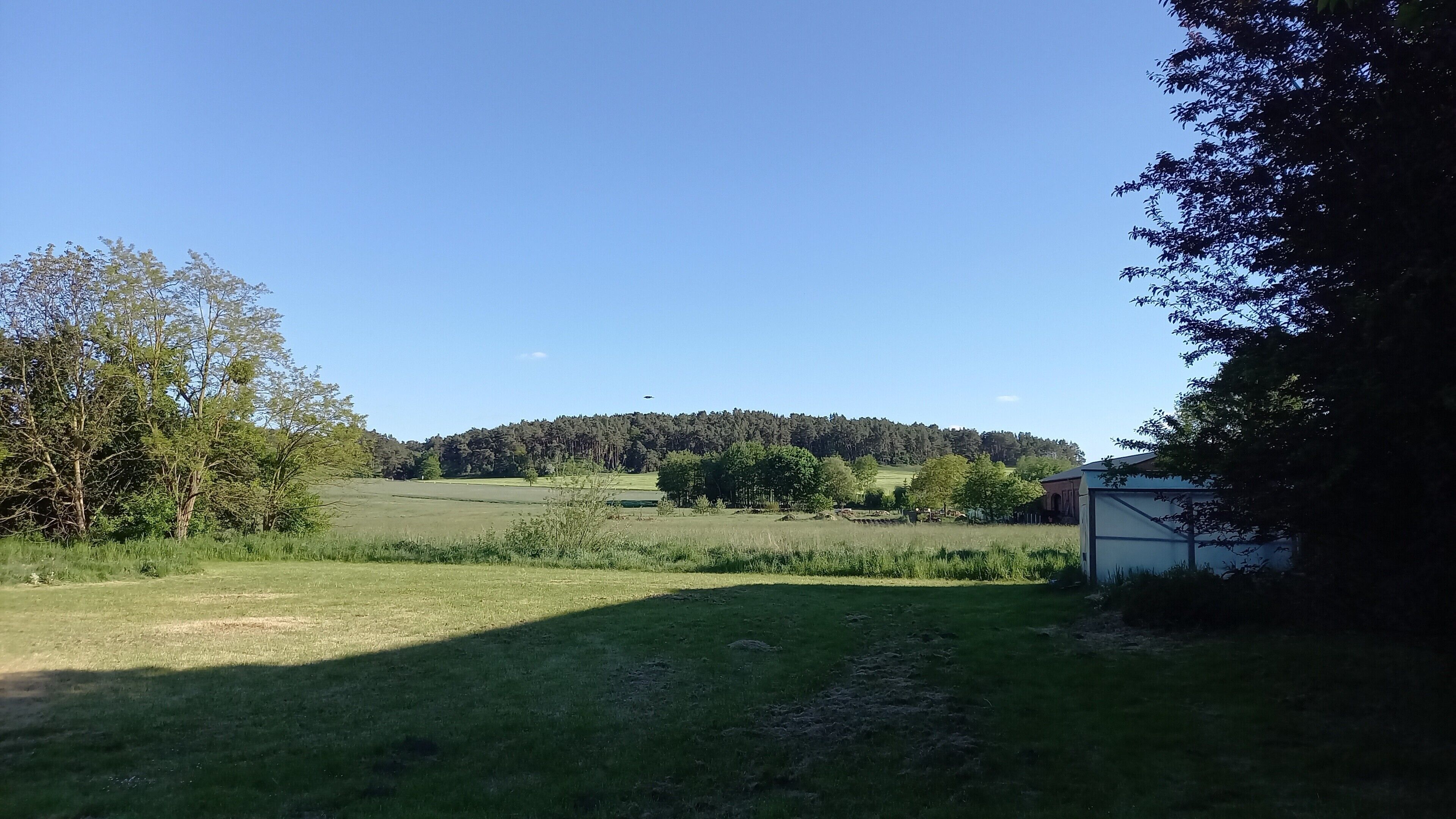 Ländliche Landschaft mit blauem Himmel, einem Hügel in der Ferne und einem kleinen Gebäude rechts.