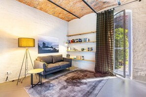 En moderne stue med sofa, gulvlampe, bogreol og et stort vindue med gardiner.