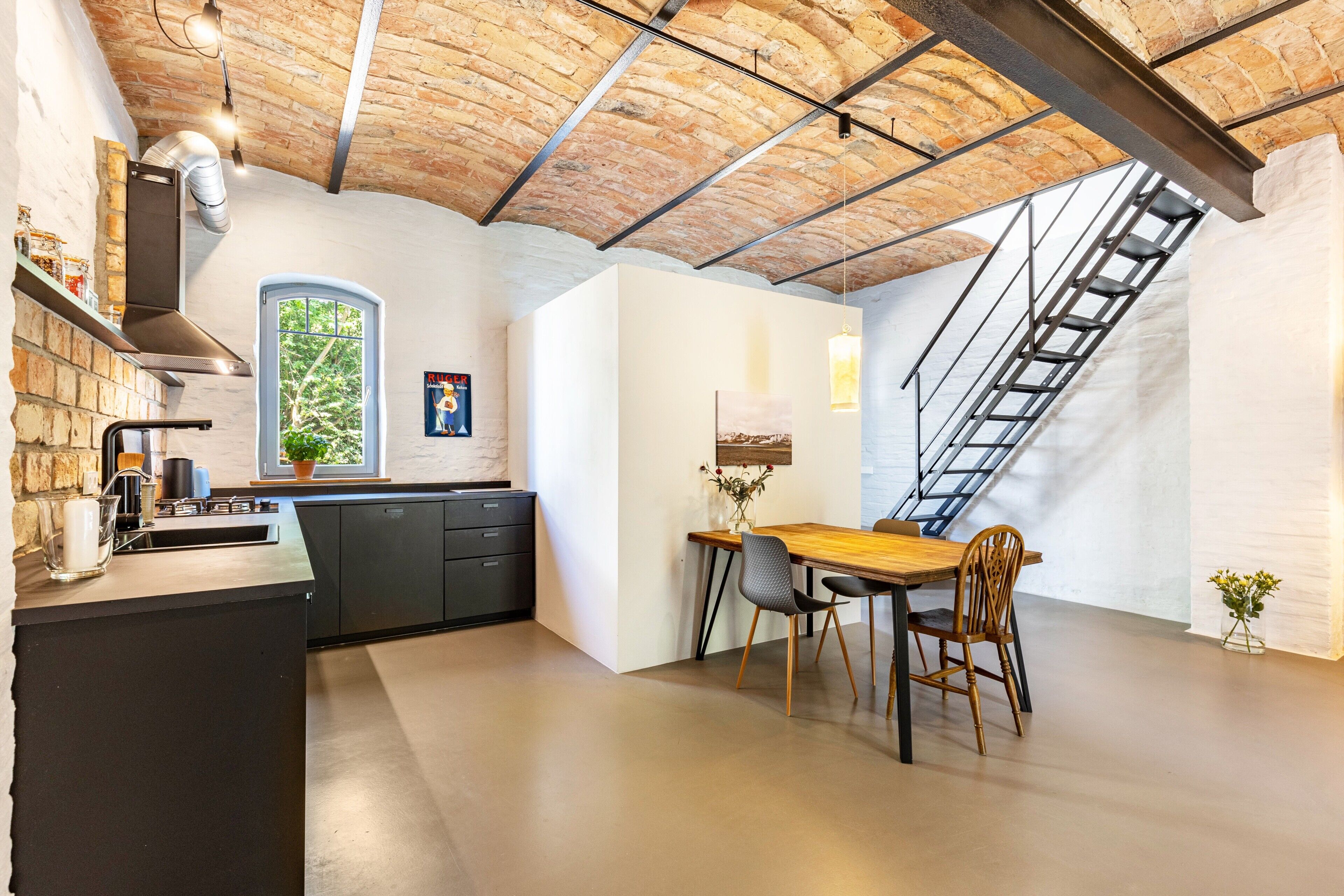 A kitchen with a brick ceiling, a window, a dining table with chairs, and a staircase.