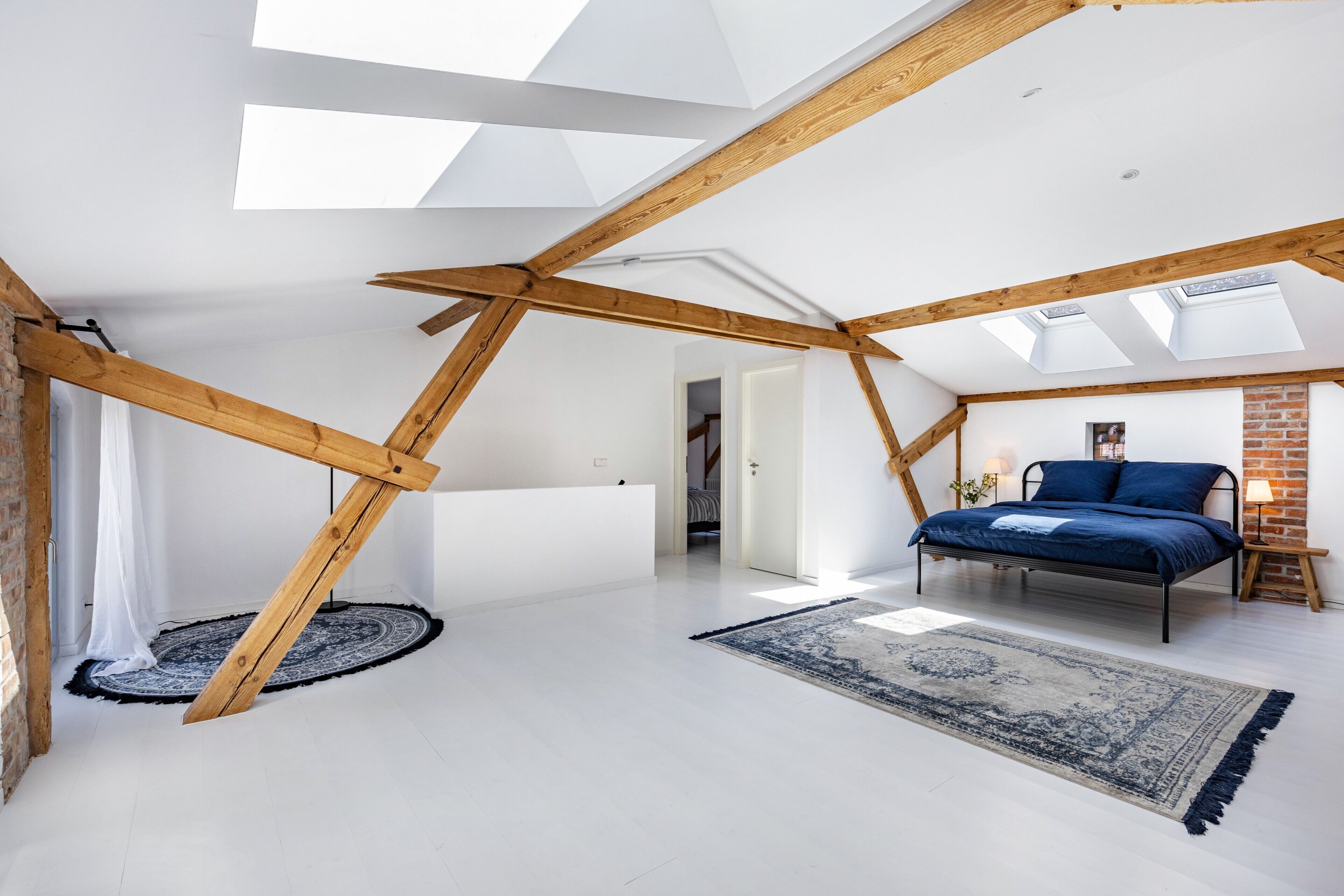 A loft bedroom with exposed wooden beams, a skylight, a bed, and a patterned rug.