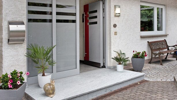 A modern building entrance with a sliding glass door, potted plants, a bench, and a small window.
