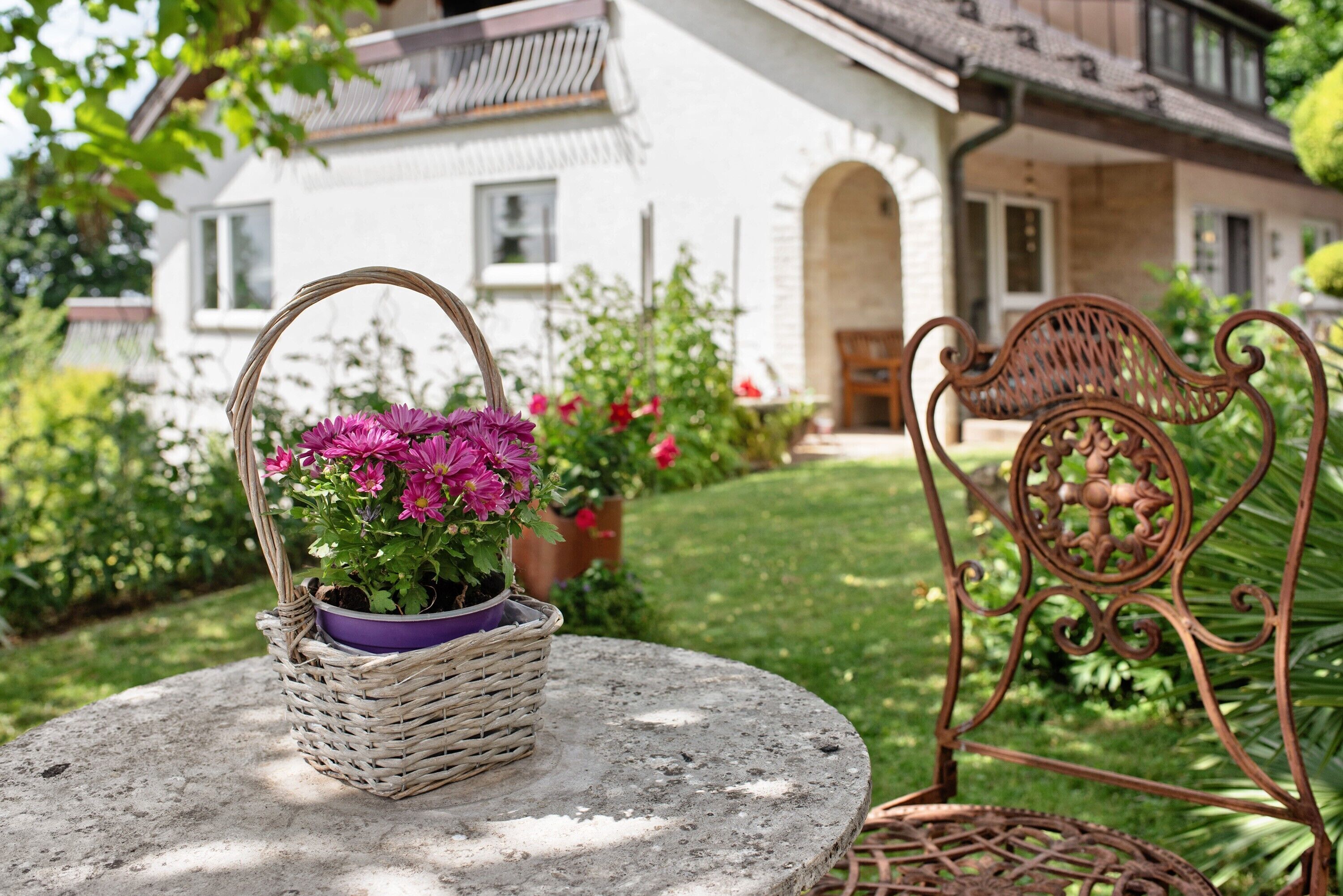 Una cesta de mimbre con flores sobre una mesa de piedra, una silla de hierro forjado y una casa al fondo.