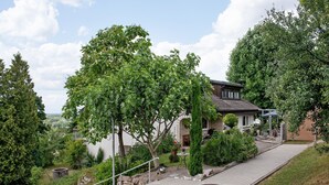 A house with a paved driveway, a small garden with a tree, and a view of the surrounding landscape.