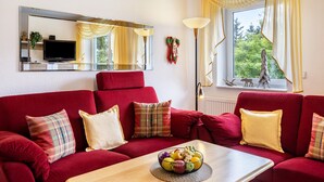 Un sofá rojo con varios cojines, una mesa de centro de madera con un frutero, un espejo grande y una ventana con cortinas.