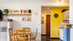 Un comedor con una mesa y sillas de madera, una estantería con libros y objetos decorativos, un reloj y una zona de cocina con una pared amarilla y una puerta que da a otra habitación.