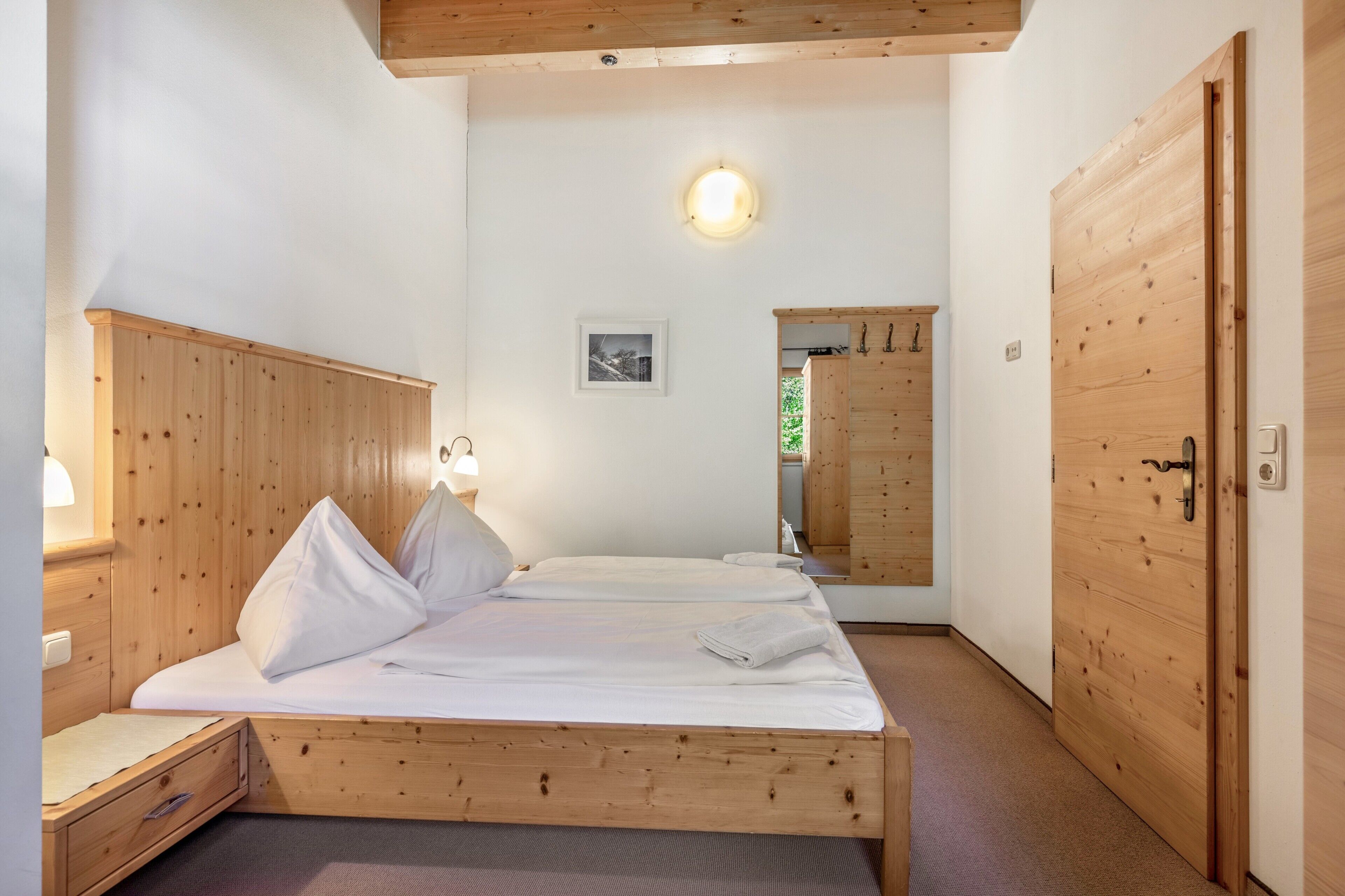 A wooden bed with a headboard, a bedside table with a lamp, a framed picture on the wall, and a wooden wardrobe.