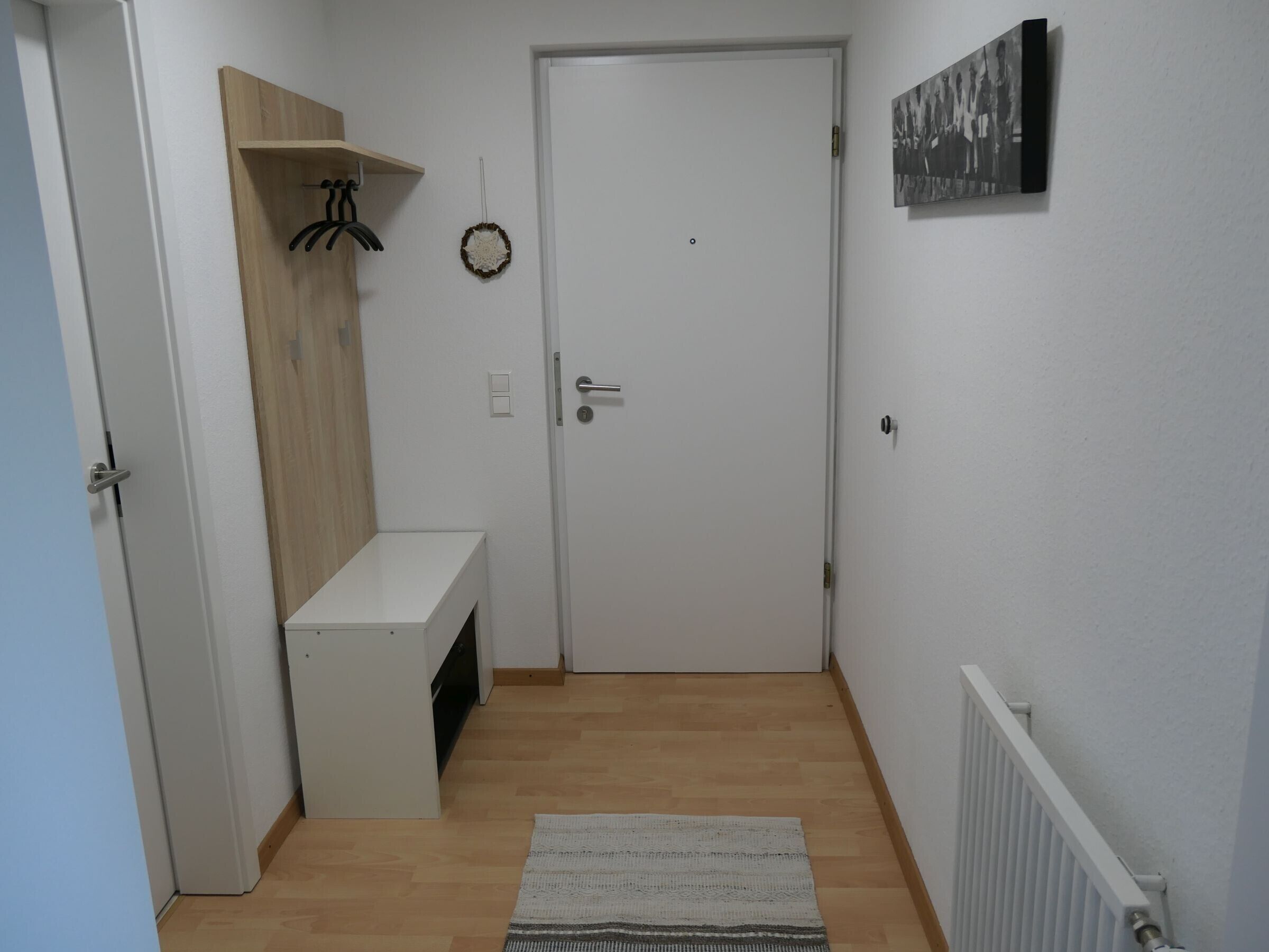 Un couloir avec une porte blanche, un petit banc blanc, une étagère en bois, un interrupteur fixé au mur et un radiateur.