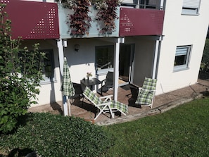 Ein überdachter Sitzbereich mit Stühlen und einem Sonnenschirm, ein weißes Gebäude mit Balkon und ein Heckenstreifen im Vordergrund.