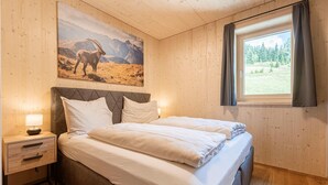 Un dormitorio con cama, mesitas de noche, lámpara, ventana con cortinas y un cuadro de un paisaje montañoso en la pared.