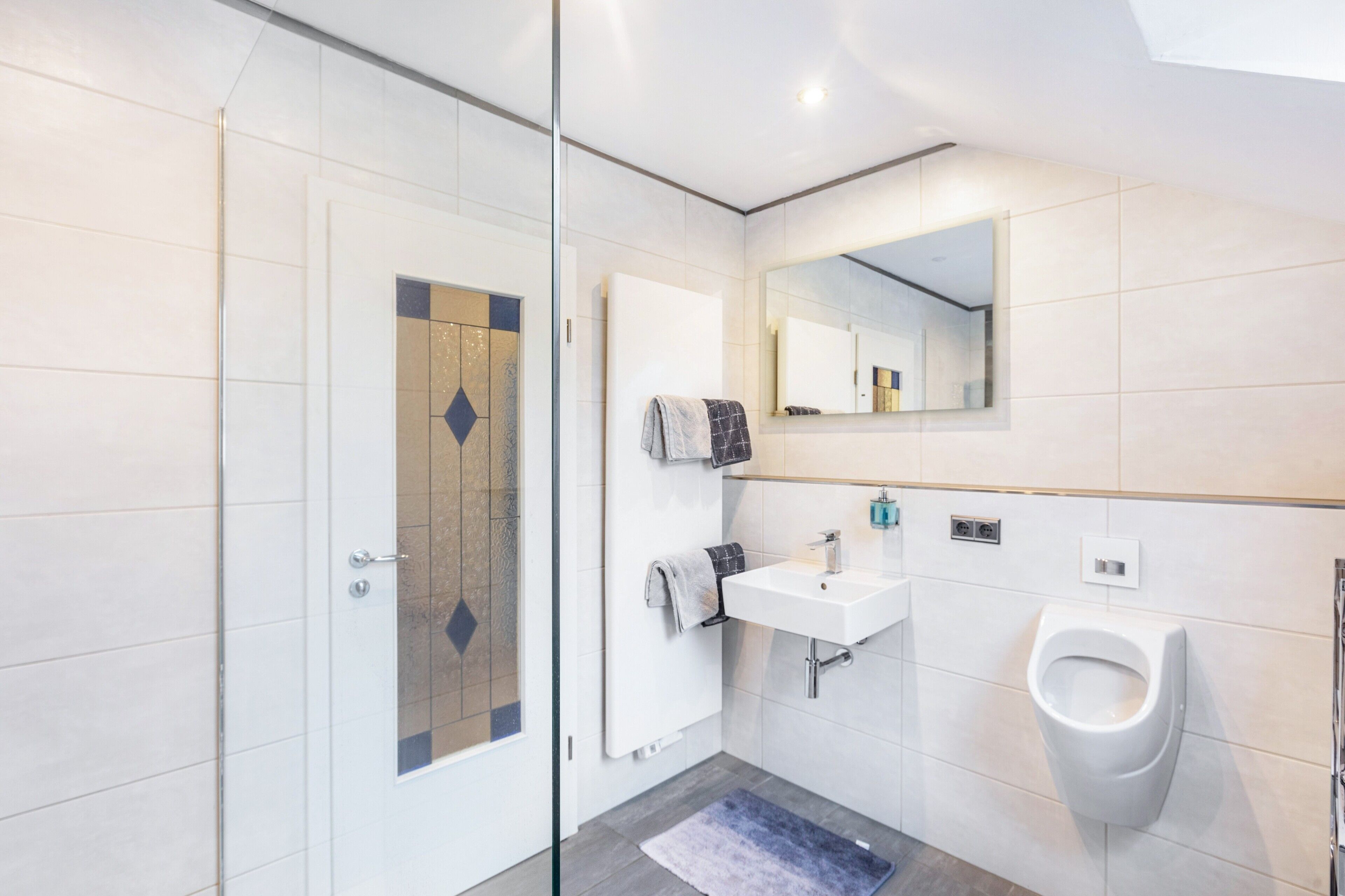 Et moderne badeværelse med en glasbrusekabine, en hvid vask, en pissoir og et spejl over vasken.
